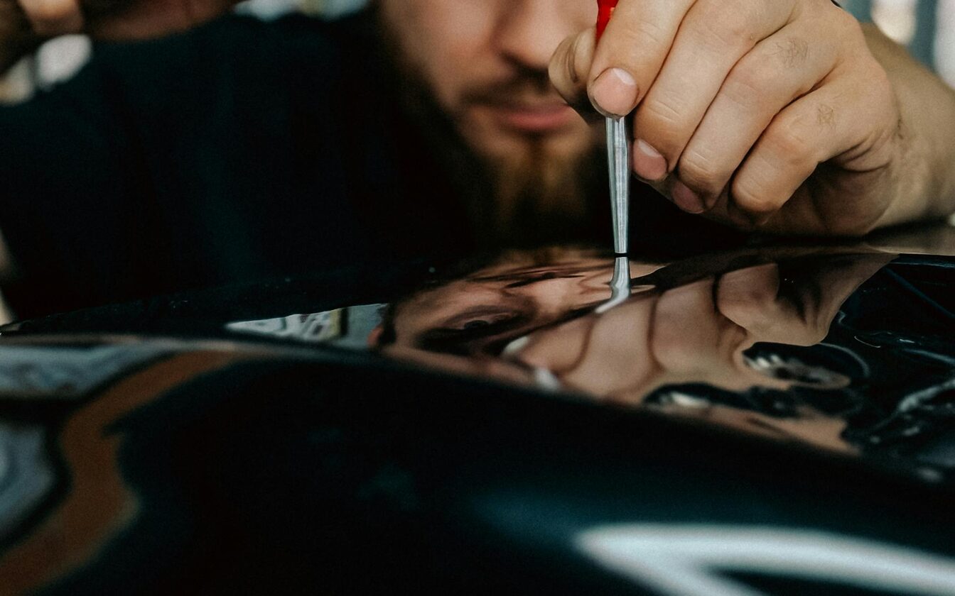 Paintless dent repair technician pressing a calibrated steel rod against a dark car's glossy panel, the reflection revealing the dents, inside a Dent Barn shop bay